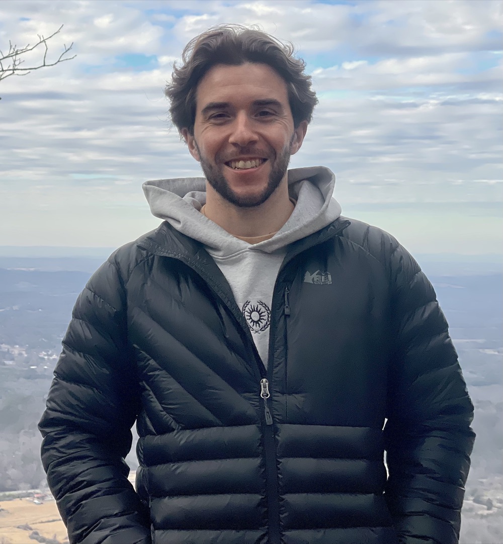Jared Krupp smiling on a mountain summit, with a valley and clouds behind him